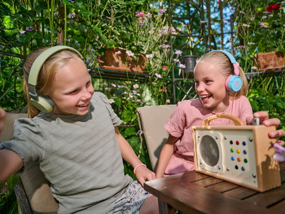 hörbert is sitting on a garden table, and two girls are listening with Bluetooth headphones
