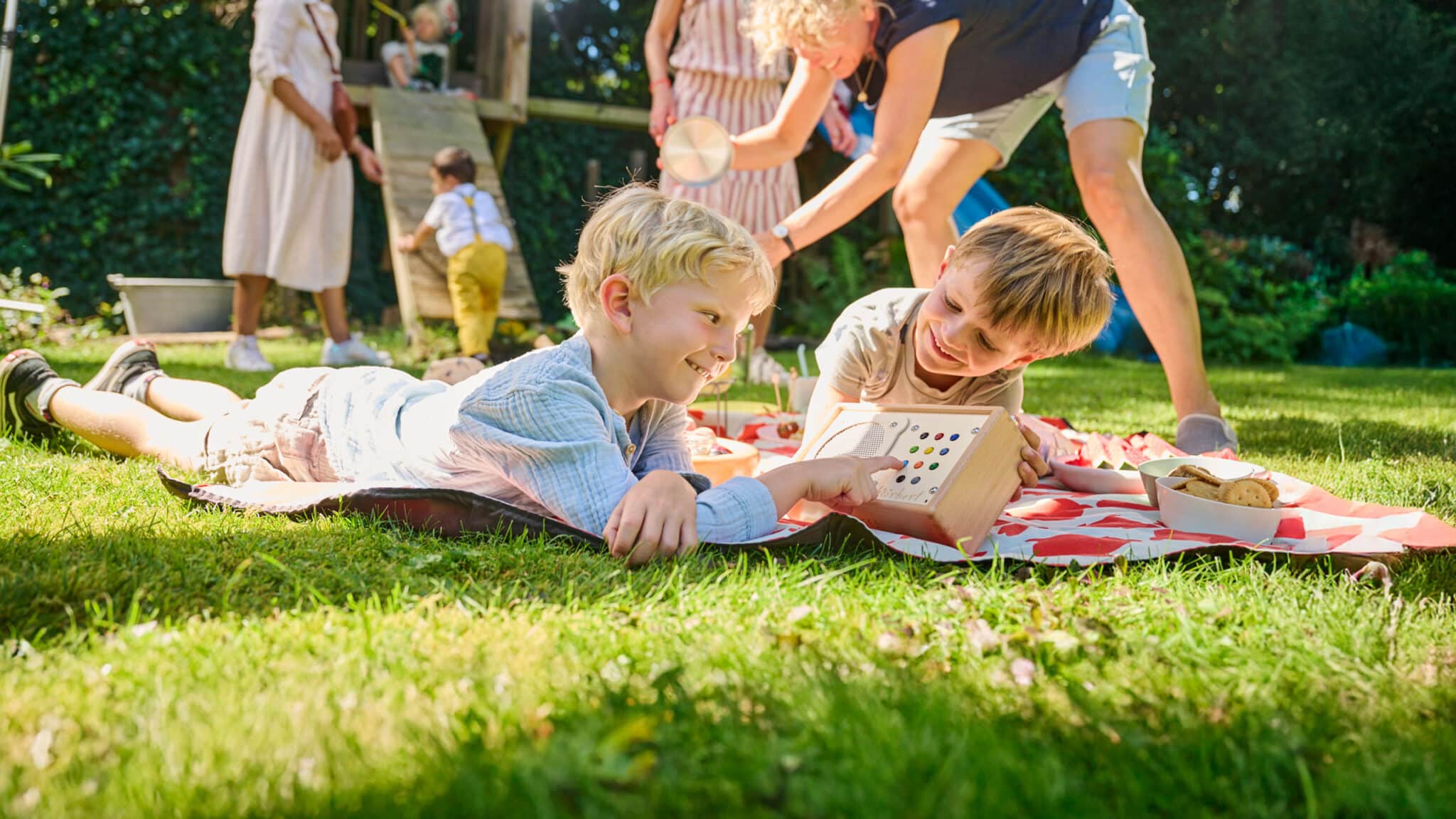 Two boys listening to hörbert on the picnic blanket while other children play in the background
