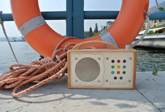 This picture shows a Hörbert music player with nine playlists, standing on a jetty by a body of water. In the background, you can see an orange and white striped lifebuoy and some ropes.